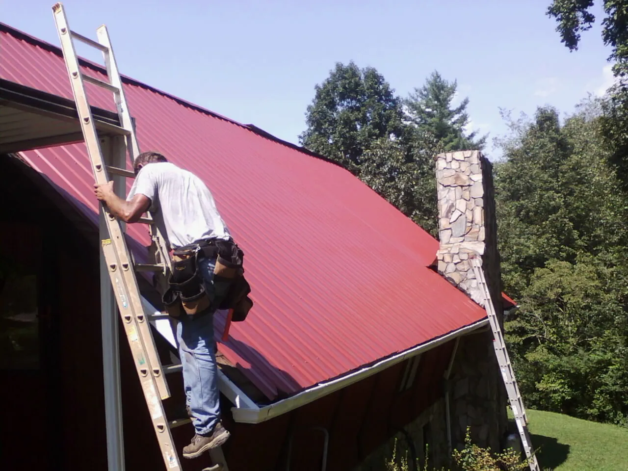 Roofer on a ladder beside a red metal roof and stone chimney — Soffit & Fascia Repair by Roofing Brothers in Blaine